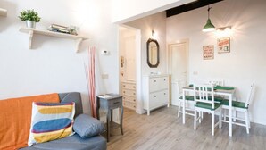 A living room with a grey sofa, a small table, a white cabinet, and a dining area with white chairs and a green tablecloth. - GuestHost - Functional 35sqm apartment, well furnished, that can accommodate up to 4 people. Located on the third floor, without lift, of an historic Florentine building few steps away from Palazzo Medici Riccardi and 3 minutes on foot from the Duomo. Th (Firenze)