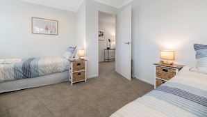 Un dormitorio con dos camas, mesitas de noche con lámparas, un cuadro enmarcado en la pared y una puerta que da a otra habitación.