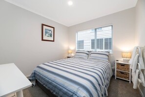 A bedroom with a bed, a bedside table, a lamp, a framed picture on the wall, and a window with blinds. - Aotahi - The Star of the Mount (Mount Maunganui)