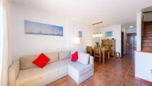 Een woonkamer met een beige bank, rode kussens, een eettafel met stoelen, een houten kast en een schilderij van zeilboten aan de muur.