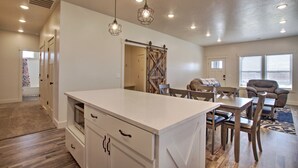 A spacious kitchen with a large island, wooden dining table, and chairs. There is a sliding barn door closet and a window with blinds. - Blue Bear Bungalow - Pool Access and only One Block From The Lake! (Garden City)