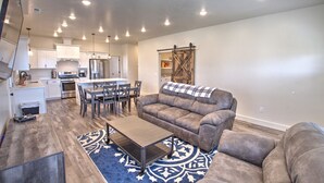 A spacious living room with a large gray sofa, a coffee table, and a patterned rug. There is a kitchen area with white cabinets and stainless steel appliances. - Blue Bear Bungalow - Pool Access and only One Block From The Lake! (Garden City)