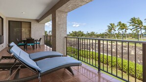A balcony with two lounge chairs, a table, and chairs, overlooking a landscaped area with palm trees and a view of the ocean.