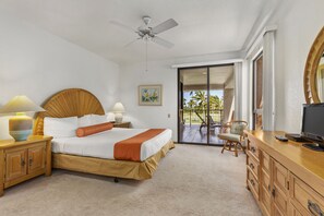 Quarto de hotel com cama, abajures, ventoinha no teto, televisão, cômoda de madeira e porta de vidro corrediça para uma varanda com vista para palmeiras.
