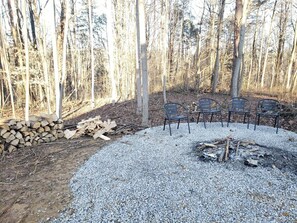 A forested area with a gravel fire pit surrounded by metal chairs.