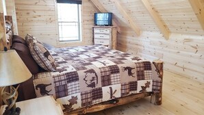 Ein Schlafzimmer einer Holzhütte mit einem Bett, einem Nachttisch, einer Lampe und einem Fenster.