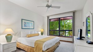 Ein Hotelzimmer mit einem Bett, Nachttischen, einem Schreibtisch, einem Deckenventilator, einem Fenster mit Blick auf Bäume und einem gerahmten Bild an der Wand.