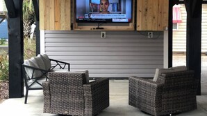 Un espace extérieur aménagé avec des sièges, abrité par un toit, comprenant deux fauteuils en osier et un téléviseur fixé à un mur en bois.