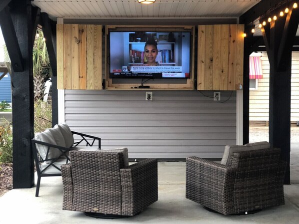 Een overdekt terras met twee rieten stoelen en een televisie die aan een houten wand is bevestigd.