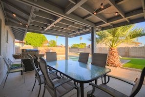 Une terrasse extérieure avec une table à plateau de verre, des chaises en métal, un barbecue et une vue sur une piscine et un palmier.