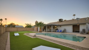 Un patio trasero con piscina, césped artificial y una zona de patio con sillas y una mesa.