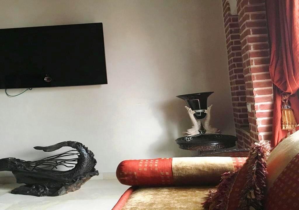 Een kamer met een wandgemonteerde flatscreen televisie, een decoratief object dat op een tak lijkt, een klein tafeltje met kandelaar, en een rood-beige gestreept kussen op een bank.