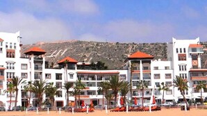 Un hotel frente a la playa con varios balcones, palmeras y montañas al fondo.