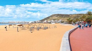 Una playa con un paseo curvo, sombrillas y una colina al fondo.
