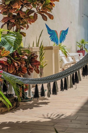 A hammock tied between plants, a wall with a wing mural, and white chairs in a courtyard.