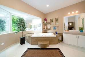 A modern bathroom with a large bathtub, a window with a view of greenery, and a built-in shelf with decorative items.