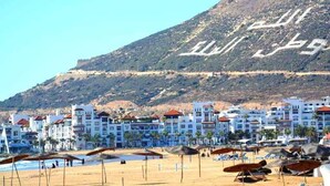 Ein Hotel an der Küste mit mehreren Etagen, Strandstühlen und einem Hügel mit arabischen Schriftzeichen.