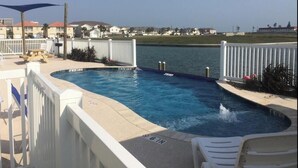 A swimming pool with a 6-inch depth marker, surrounded by a white fence, and overlooking a body of water with buildings in the background. - HUGE 4BR/3BA! Pool, Beach & Boat Slip! 4 Car Garage  (Corpus Christi)