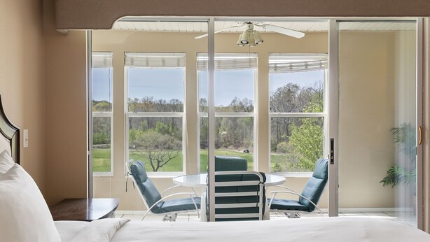 Un dormitorio con una cama grande, una mesa redonda, dos sillas y vistas a un paisaje verde a través de ventanas amplias.