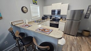Cucina con mobili bianchi, frigorifero in acciaio inossidabile, forno a microonde e macchina per il caffè. Sono presenti due sgabelli da bar e un cartello con su scritto 'BEACH HOUSE Where memories are made'.