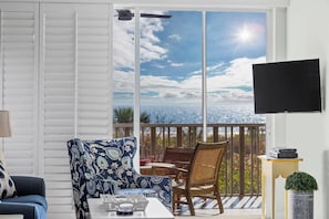 A room with a sofa, a patterned armchair, a table, a TV mounted on the wall, and a balcony with a view of the sky and clouds.
