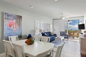 Un coin repas avec une table et des chaises blanches, un canapé bleu orné de coussins à motifs et un ventilateur de plafond.