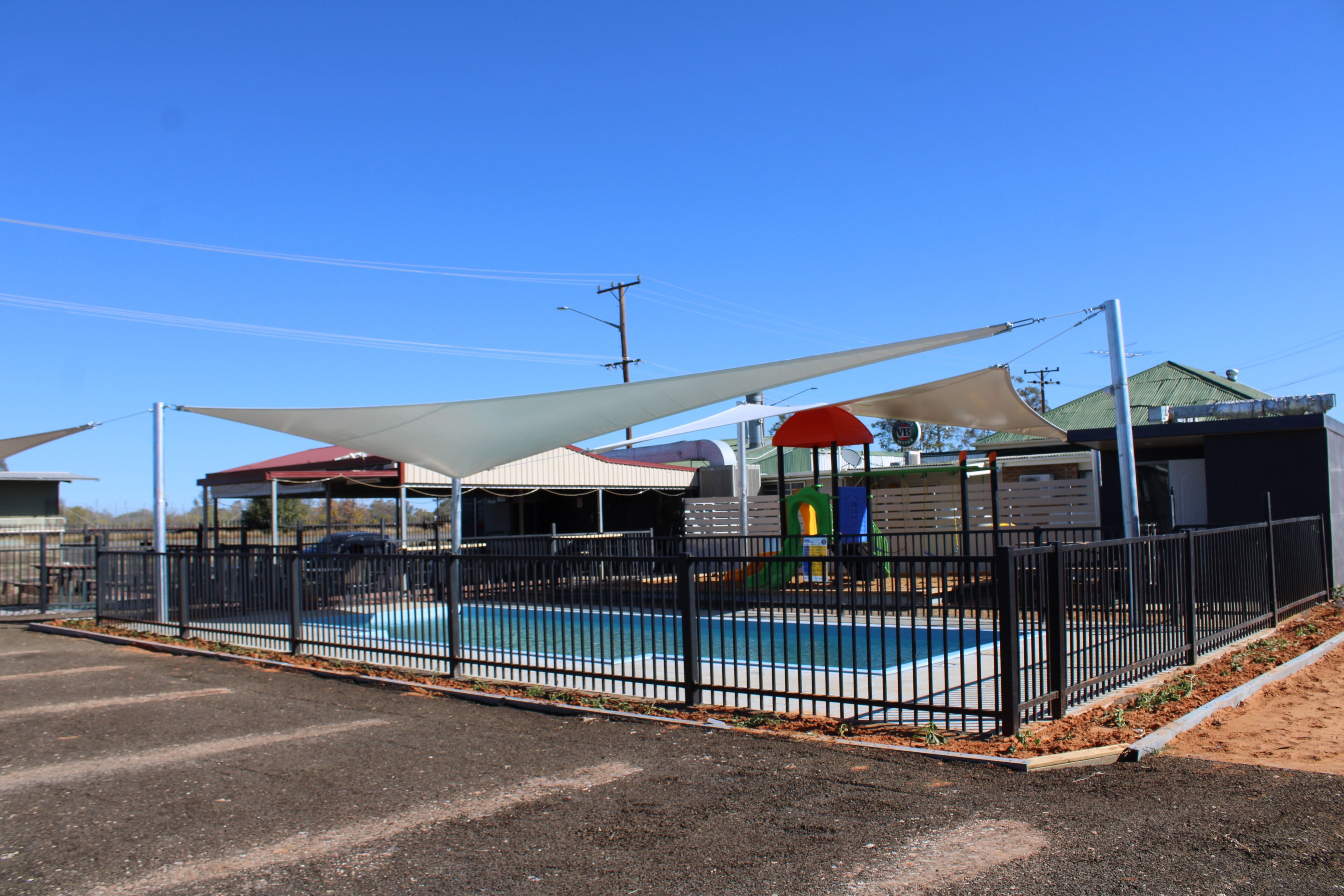 A fenced outdoor swimming pool area with a shaded canopy, a covered seating area, and a playground.