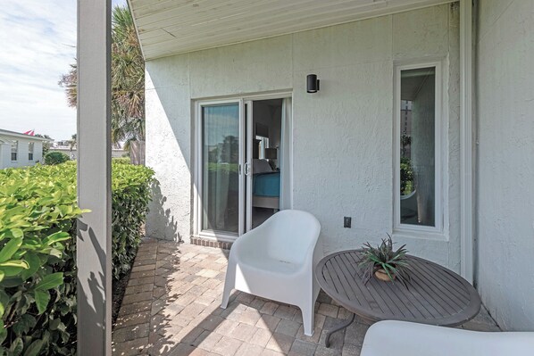 Un patio techado con una silla blanca, una pequeña mesa redonda con una planta y un ventanal corredero que da a una habitación.