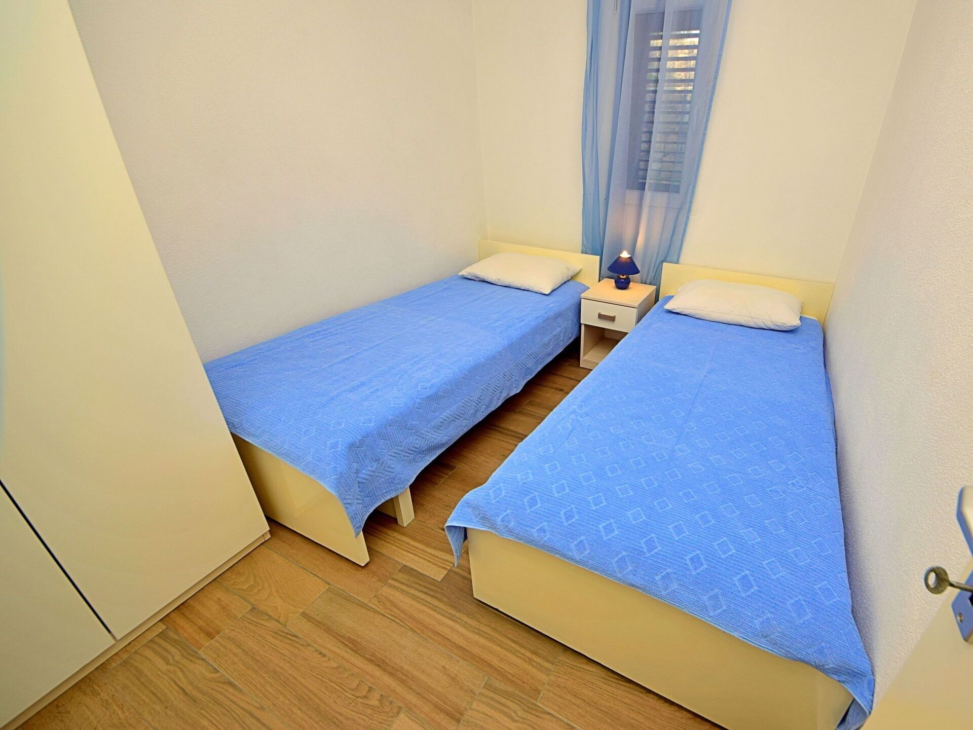 Une petite chambre propre, avec deux lits simples, une table de chevet avec une lampe et une fenêtre ornée de rideaux légers.