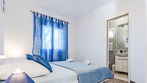 A bedroom with two beds, a nightstand with a lamp, a bathroom with a sink and mirror, and a window with blue curtains.