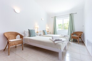 Une chambre d’hôtel avec deux lits, un coin salon avec des fauteuils, une fenêtre avec des rideaux et une lampe fixée au mur.