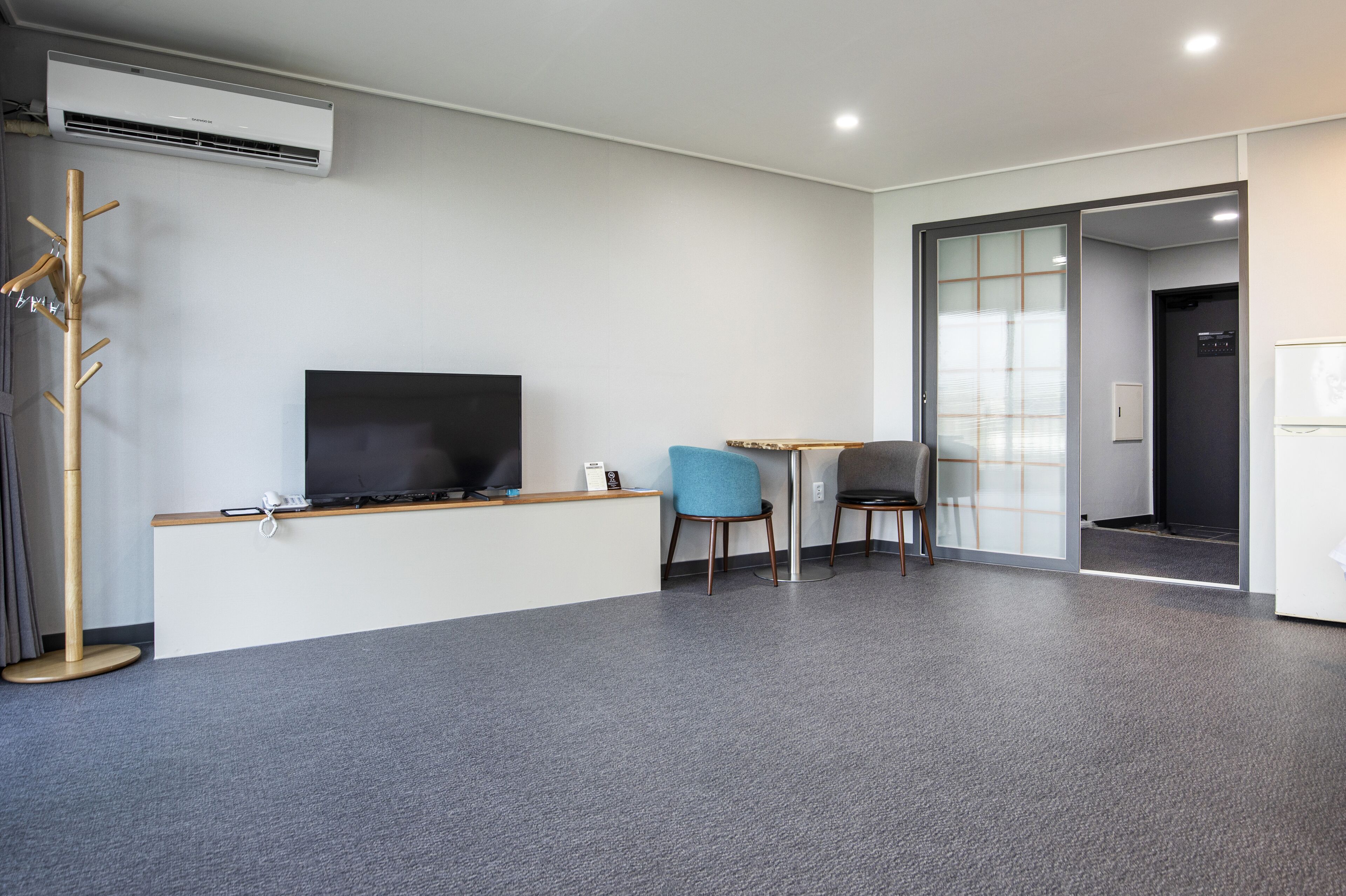 A modern hotel room with a flat-screen TV, a wooden desk, a blue chair, and a coat rack.