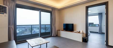 A modern hotel room with a large window, a flat-screen TV, a sofa, a small table, and a trash bin.