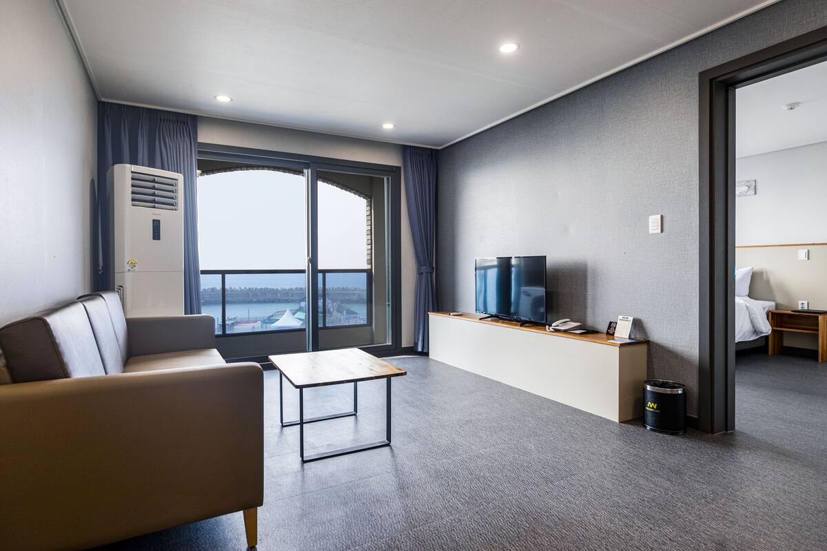 A modern hotel room with a sofa, a small table, a TV, and a view through a large window.
