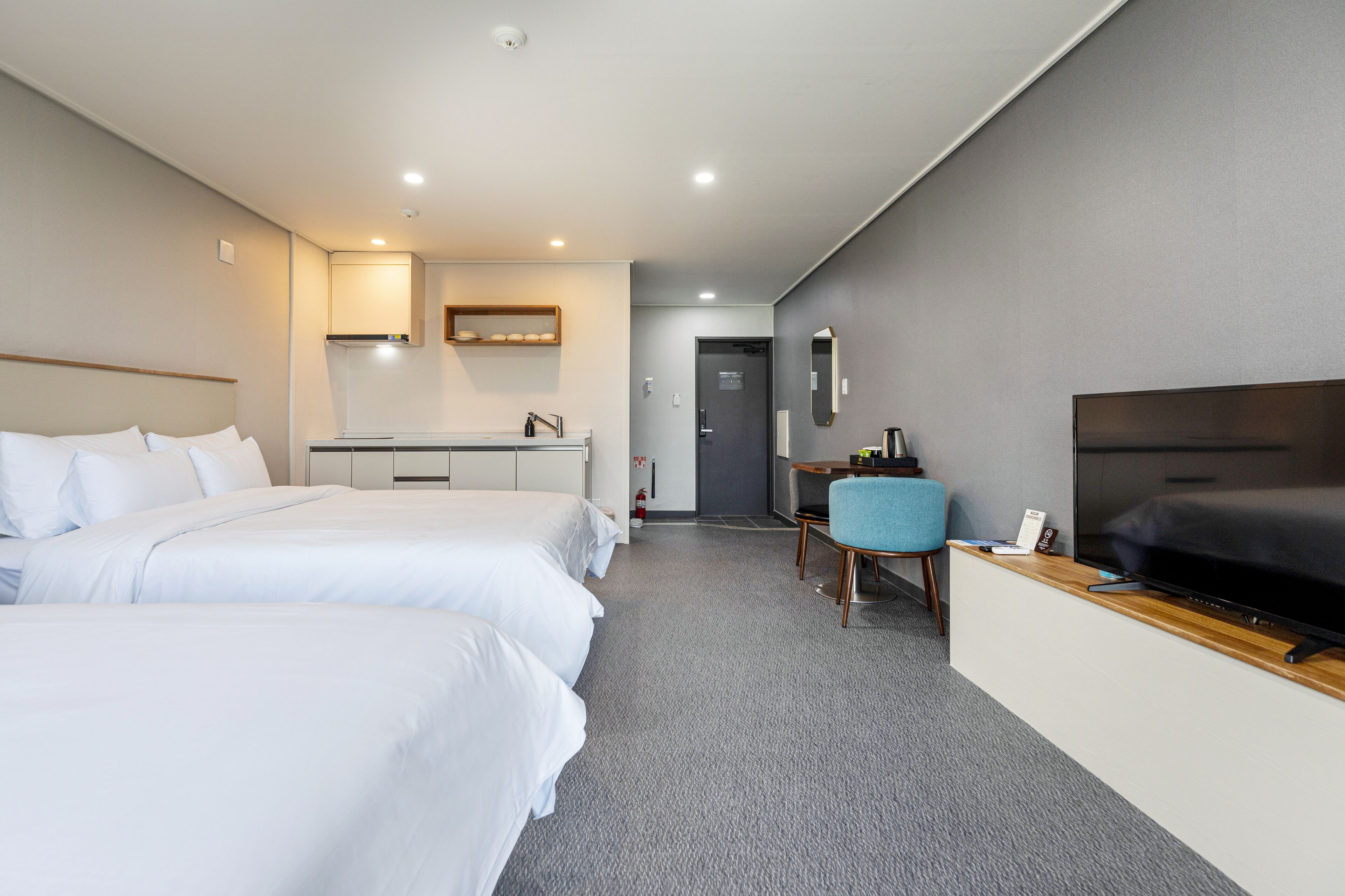 A hotel room with two beds, a desk with a chair, a flat-screen TV, and a small kitchenette.