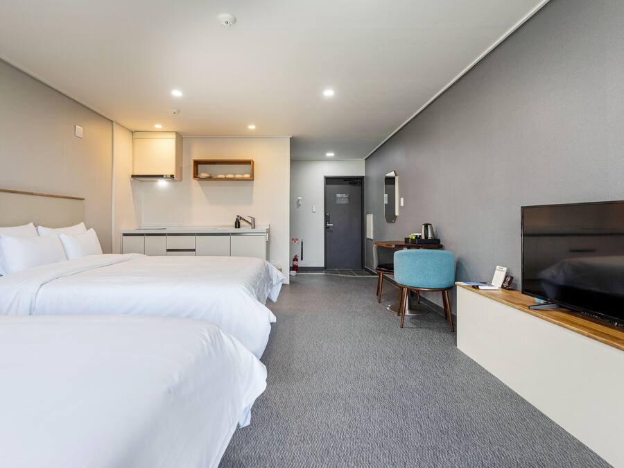 A hotel room with two beds, a desk with a chair, a flat-screen TV, and a small kitchenette.