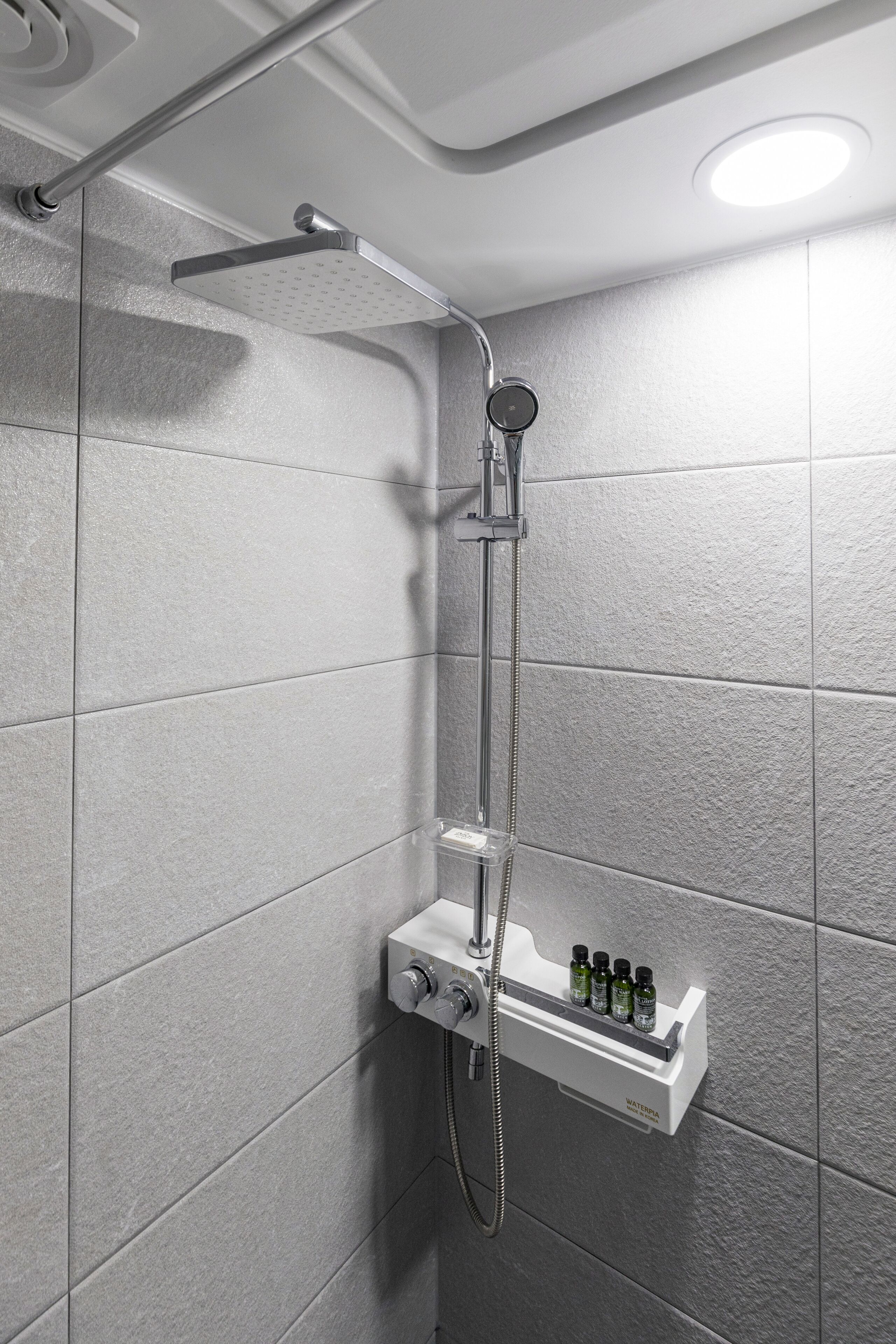 A shower with a handheld showerhead, a wall-mounted control panel, and a shelf with toiletries.