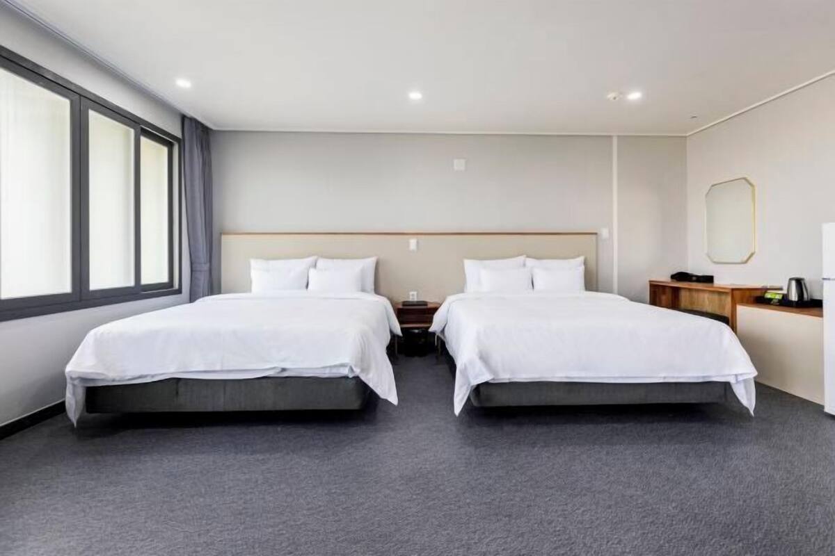 A hotel room with two beds, a large window, a wooden nightstand, and a mirror on the wall.