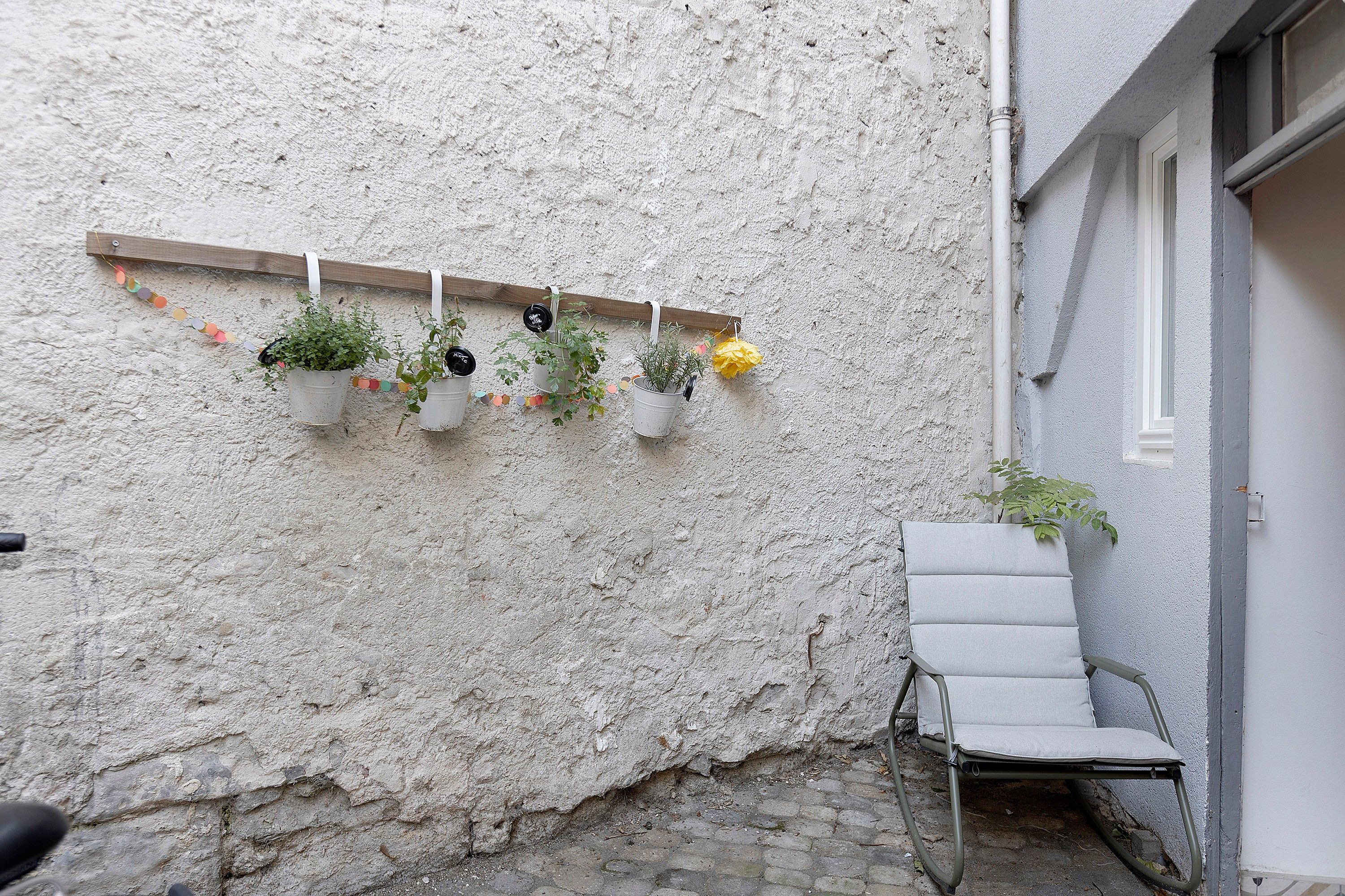 Un mur blanc à texture, agrémenté d’une étagère en bois où sont disposées des plantes en pot, et une chaise en métal.