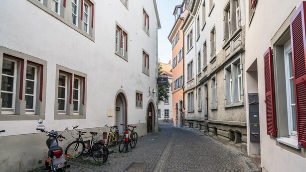 Eine enge Kopfsteinpflasterstraße, gesäumt von Gebäuden mit rundbogigen Portalen und roten Fensterläden.
