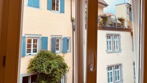 Blick durch eine offene Tür auf ein Gebäude mit zahlreichen Fenstern, einige mit Rollläden, und einen Balkon mit Pflanzen.