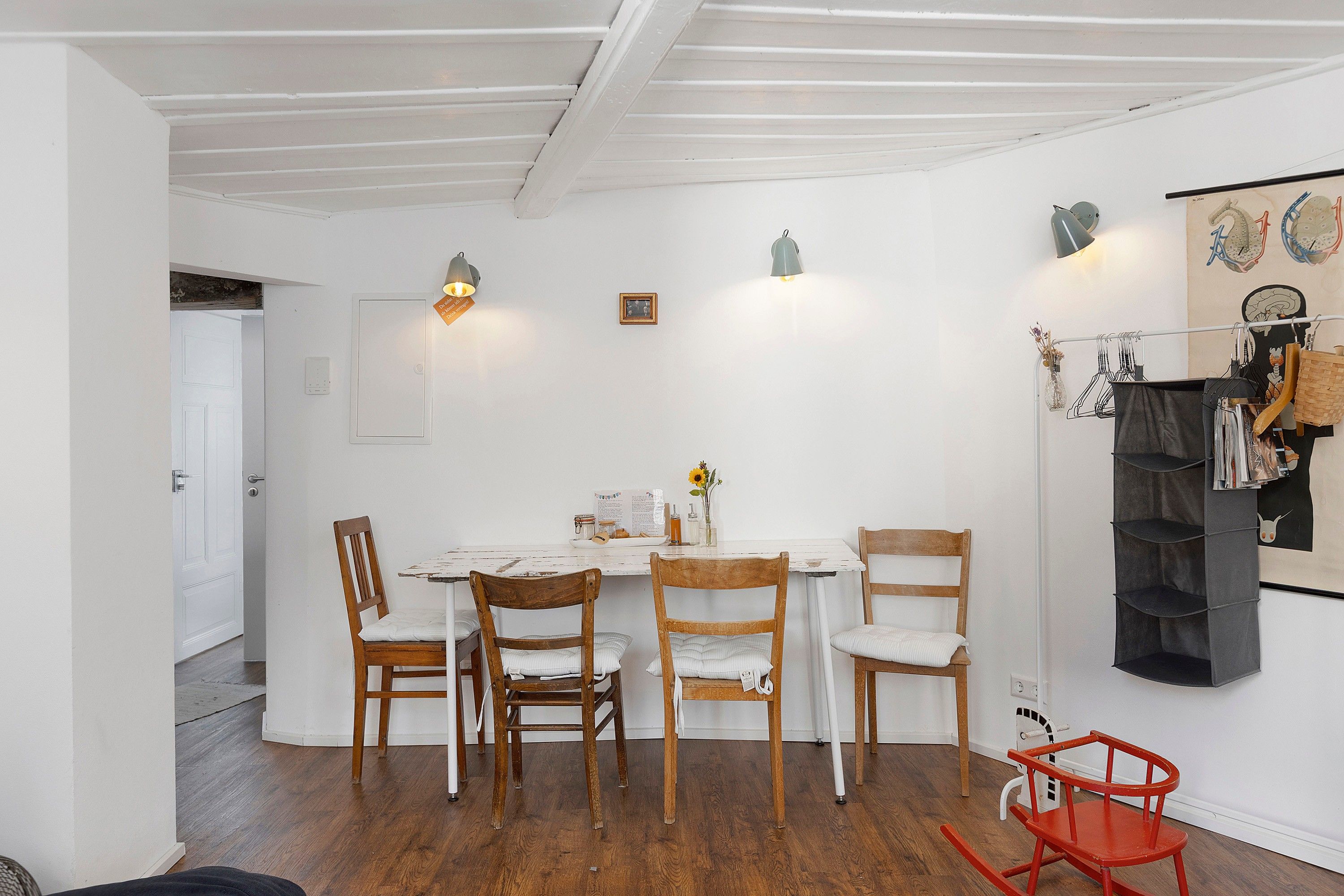 Uma sala de jantar com uma mesa branca, quatro cadeiras de madeira, uma poltrona de balanço vermelha e uma estante fixada na parede com diversos objetos.