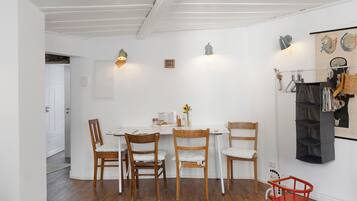Uma sala de jantar com uma mesa branca, quatro cadeiras de madeira, uma poltrona de balanço vermelha e uma estante fixada na parede com diversos objetos.
