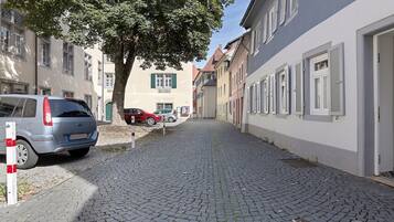Une rue pavée bordée de bâtiments traditionnels, de voitures garées et d’un arbre qui offre de l’ombre.