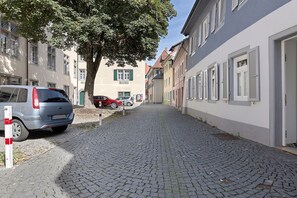 Eine Kopfsteinpflasterstraße gesäumt von traditionellen Gebäuden, parkenden Autos und einem Baum, der Schatten spendet.