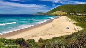 Une plage aux eaux d’un bleu limpide, bordée d’un rivage sablonneux, avec des collines verdoyantes en arrière-plan.