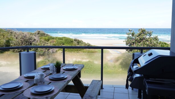 Une table en bois, dressée avec des assiettes et des verres, donnant sur une plage et l’océan.