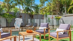 Ein Sitzbereich im Freien mit Holzmöbeln und einem Glastisch, umgeben von einem Pool und üppiger Bepflanzung.