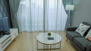 A modern living room with a flat-screen TV, a round marble coffee table with a remote control, a grey sofa with decorative pillows, and sheer curtains. - Classy 2BR Apartment at Menteng Park with City View (Jakarta)