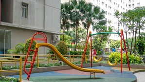 A playground with a yellow slide and red and yellow climbing structures, surrounded by greenery and a building in the background.